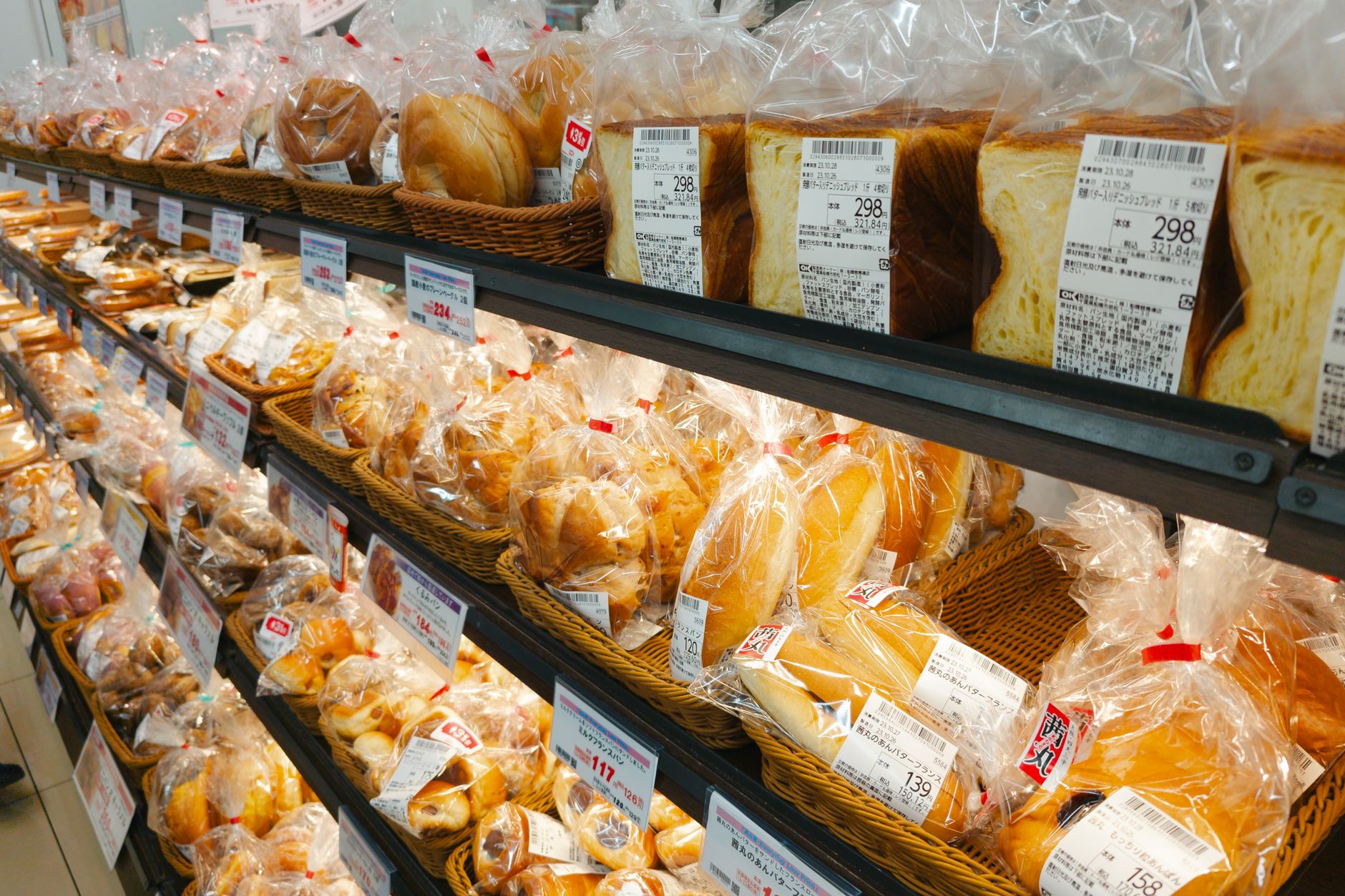 パン売り場の棚に商品ごとの値札が並ぶ店内。価格表示を確認しながら商品を選ぶ場面