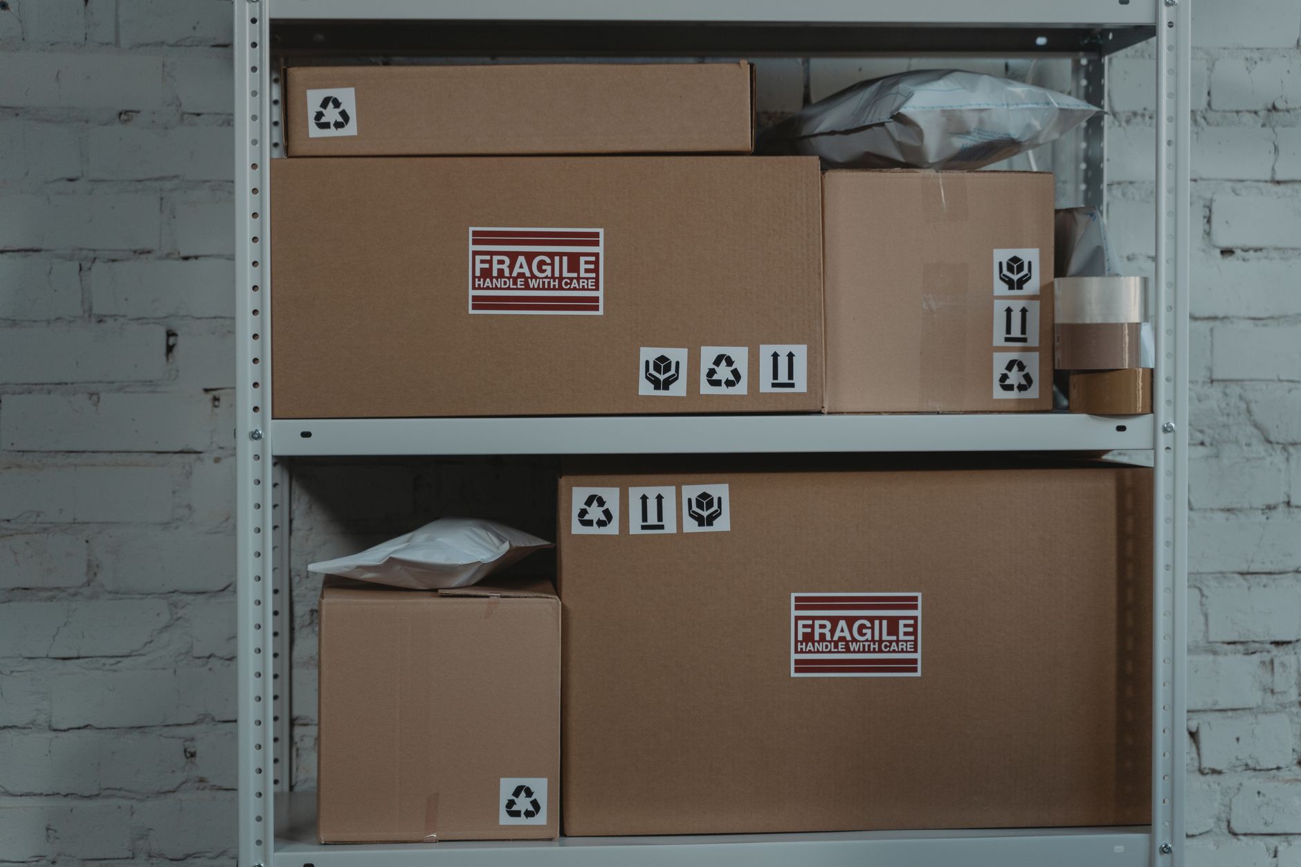 「FRAGILE」のラベルと取り扱い表示を貼った輸送用の段ボール箱を、白い倉庫のスチール棚に並べた様子